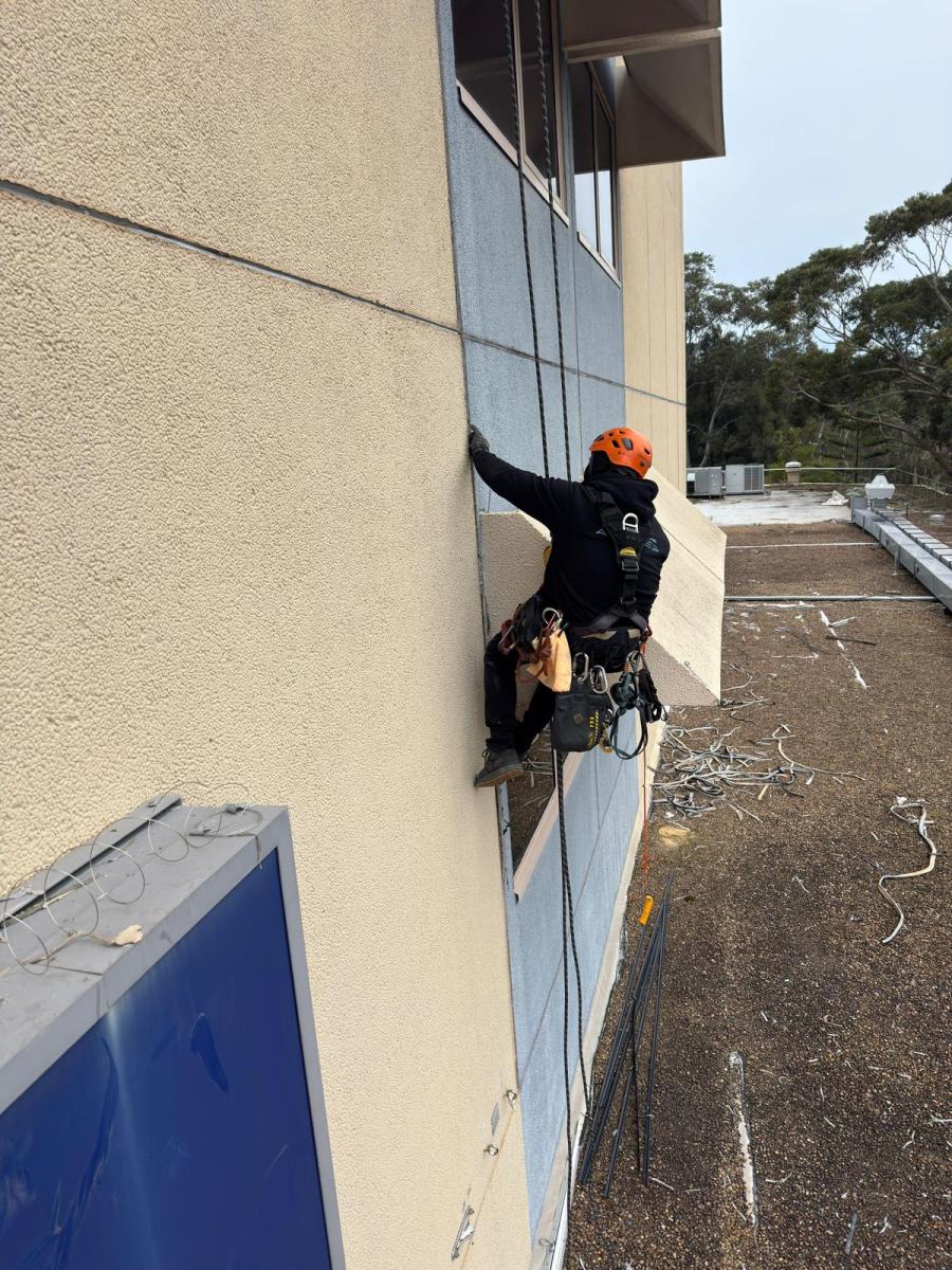 Rope access technician in orange helmet performing caulking work on commercial building facade using abseiling equipment