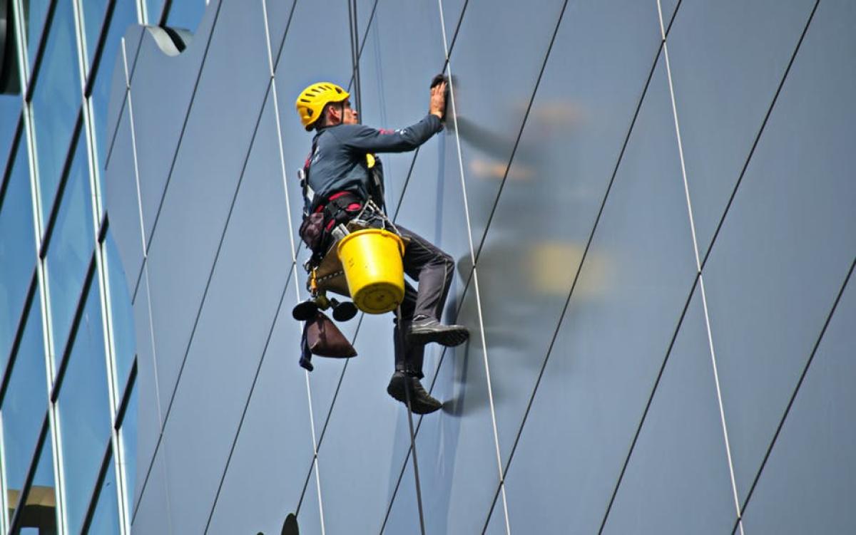 Rope access technician in yellow helmet cleaning modern building facade using professional abseiling equipment