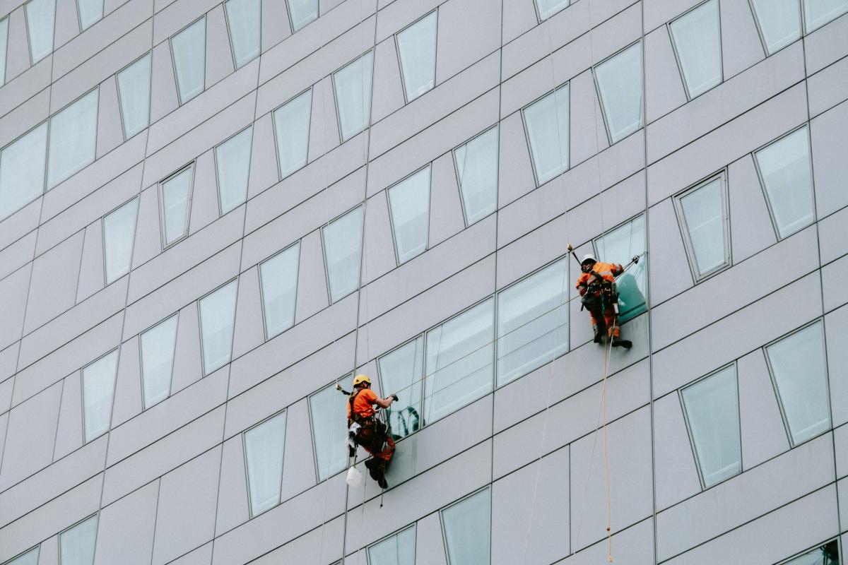 Two rope access technicians in orange safety gear cleaning windows on a modern glass office building facade
