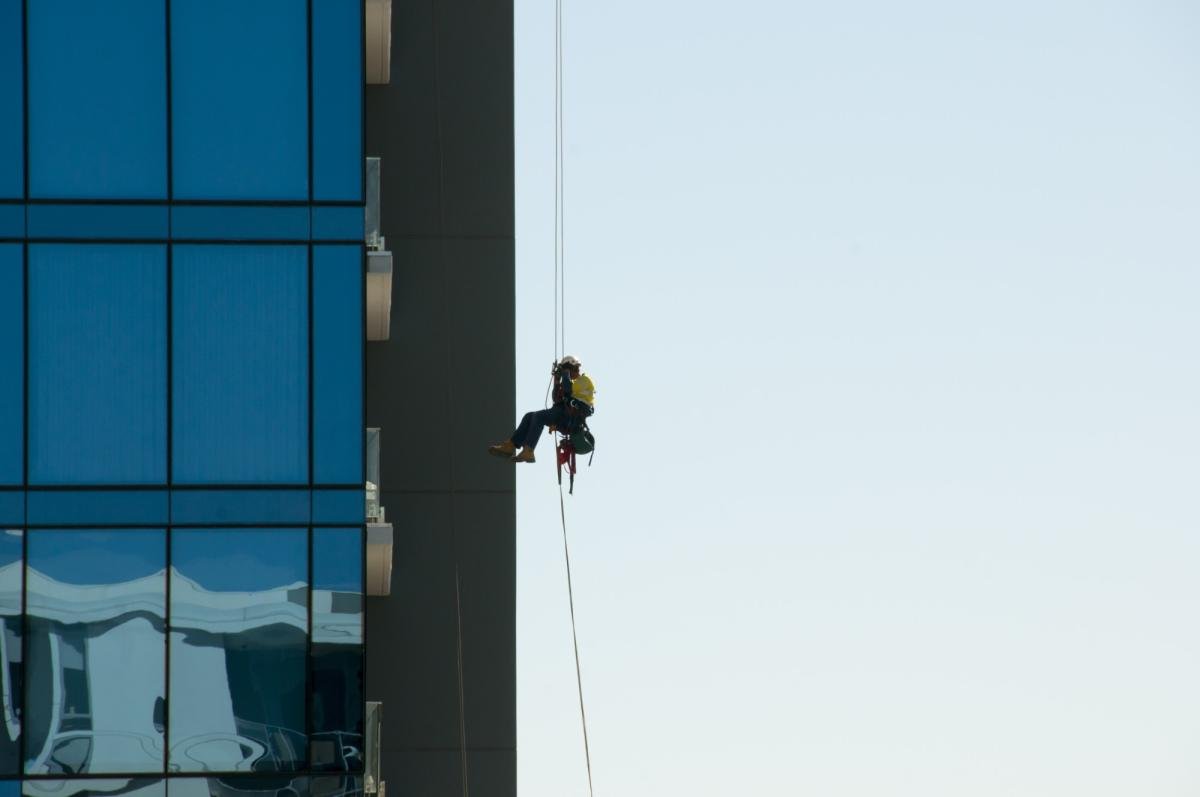 Professional rope access technician in yellow safety gear cleaning windows on modern high-rise building facade