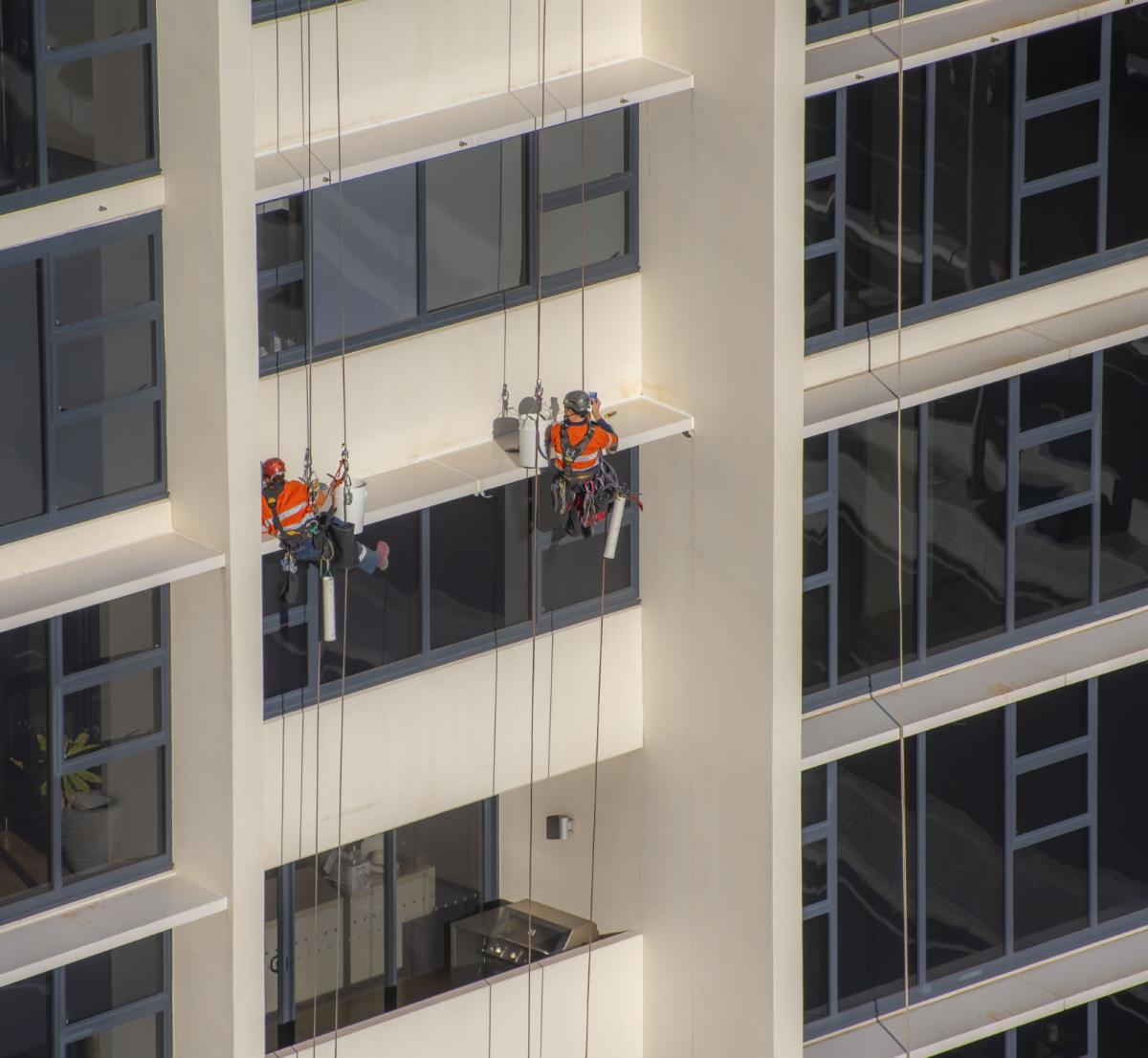 Two rope access technicians in orange high-vis jackets cleaning windows on modern high-rise building facade