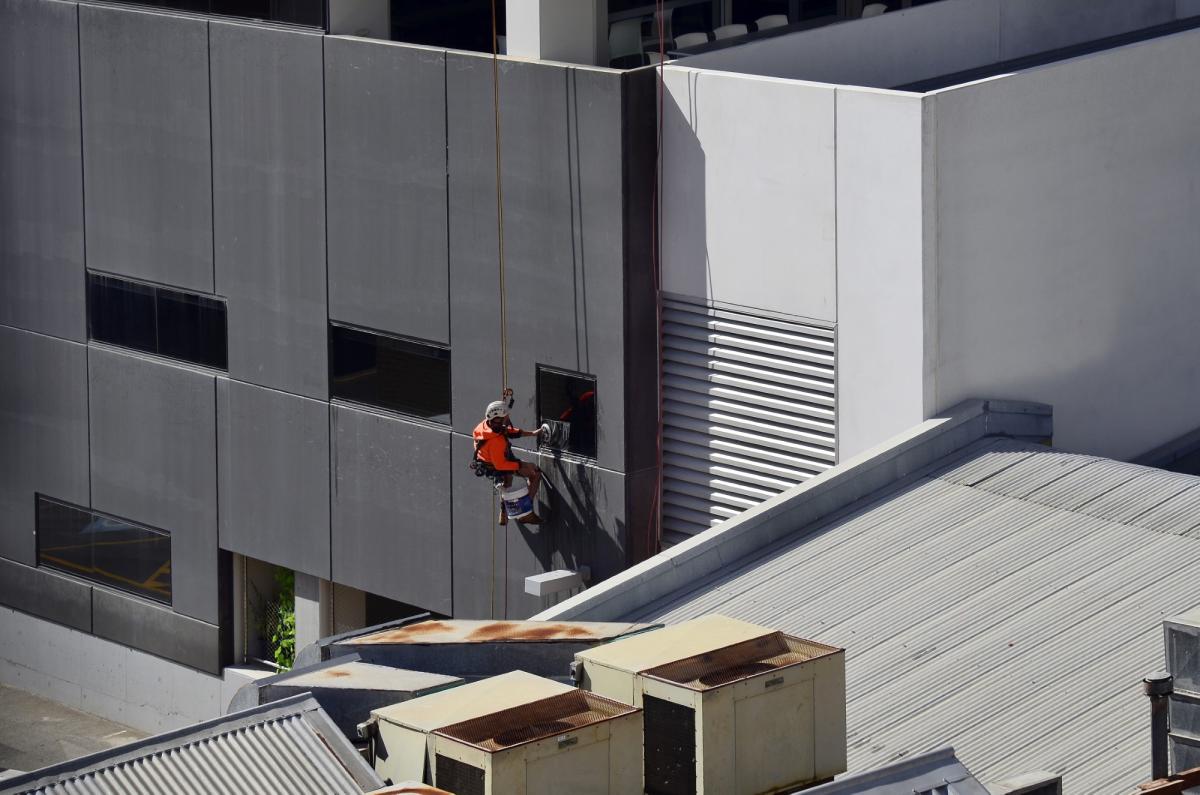 Rope access technician in orange safety gear abseiling down modern commercial building facade for maintenance work