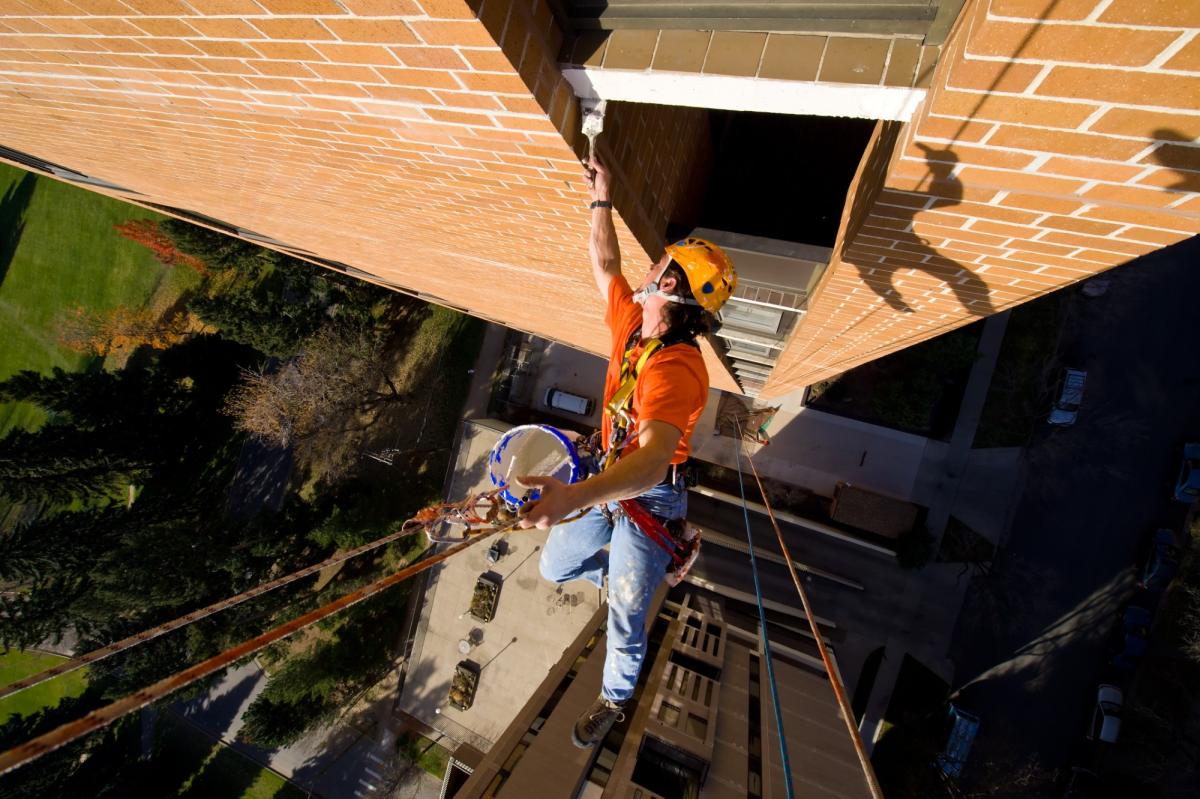 Rope access technician in orange safety gear and helmet performing building maintenance work at height